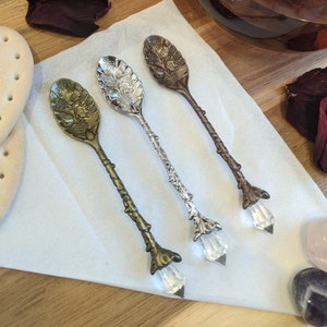 May include: Three ornate tea spoons with crystal handles. The spoons are silver, gold, and bronze with floral designs. The spoons are on a white surface with a few rose petals and crystals.