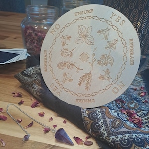May include: A wooden pendulum board with the words "Yes", "No", "Maybe", "Rephrase", and "Unsure" printed around the edge. The board is decorated with various symbols and designs, including a sun, a moon, and a hand.