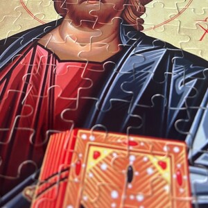 May include: A jigsaw puzzle depicting a religious icon of Jesus Christ holding a book. The icon is painted in a traditional style with a red and blue robe and a gold halo. The puzzle pieces are visible in the image.