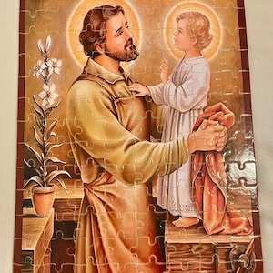 May include: A religious jigsaw puzzle featuring a painting of Saint Joseph holding a young Jesus. The image is set in a workshop with a potted plant in the background.