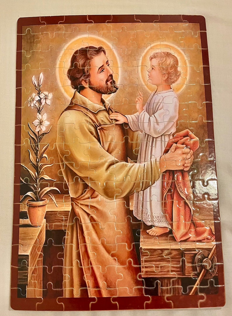 May include: A religious jigsaw puzzle featuring a painting of Saint Joseph holding a young Jesus. The image is set in a workshop with a potted plant in the background.