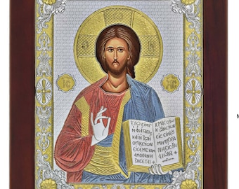 May include: A silver and gold framed icon of Jesus Christ holding a book with Greek text. The icon is on a dark wood background.