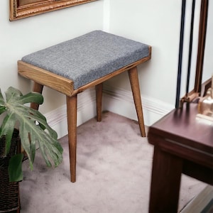 May include: A gray upholstered stool with wooden legs. The stool is in a corner of a room with a light gray carpet.