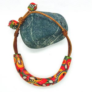 Op de afbeelding: Een kleurrijke ketting met een patroon keramische buisvormige hanger in rood, geel en wit. De ketting heeft een bruin leren koord en twee decoratieve keramische kralen. De kralen en hanger hebben een vergelijkbaar ontwerp.