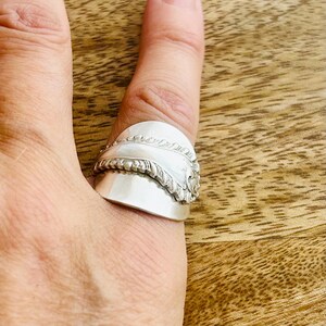 May include: A silver ring crafted from a repurposed spoon, featuring a wide, polished band with a prominent embossed decorative pattern along the centre. The metal has a bright, reflective finish and wraps around the finger with a smooth, curved design.
