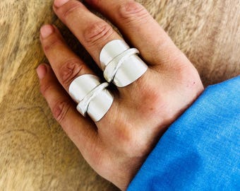 Könnte beinhalten: Zwei breite, polierte Silberringe am Mittel- und Ringfinger. Jeder Ring hat eine glatte, reflektierende Oberfläche mit einem markanten diagonalen Band, das sich um die Mitte legt und ein geschichtetes, architektonisches Aussehen erzeugt.