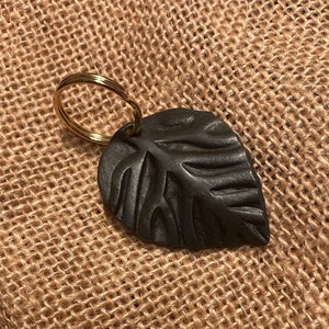 May include: A black leaf-shaped keyring with a gold metal ring. The keyring is on a brown hessian background.