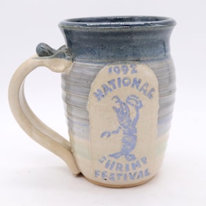 May include: Handmade ceramic mug with a blue and cream glaze. The mug features a light blue design of a shrimp and the text "1992 National Shrimp Festival". The handle is a light cream color.