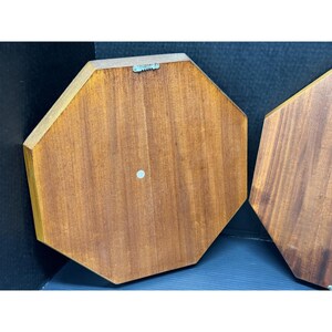 May include: Two wooden, octagonal wall plaques with a warm, reddish-brown finish. Each plaque has a small, circular, light-colored button and a metal sawtooth hanger on the back for easy mounting. The wood grain is visible.