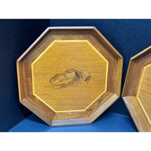 May include: Two octagonal wooden trays with a crab inlay. The trays are made of light and dark brown wood, with a lighter wood border. The crab is depicted in shades of brown and tan. The trays are likely for serving or display.