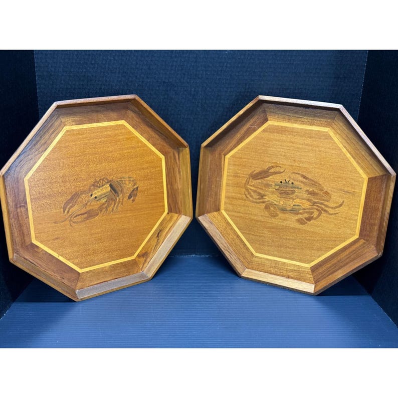 May include: Two octagonal wooden trays with a light brown finish. Each tray features a crab or lobster inlay in the center, surrounded by a lighter wood border. The trays are displayed on a blue surface.