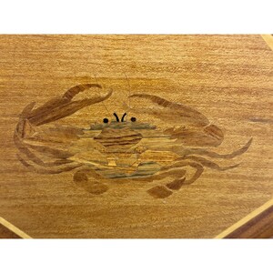 May include: Wooden inlay featuring a crab design. The crab is crafted from various wood tones, with a detailed body and claws. The background is a light-colored wood with a visible grain, and the overall design is geometric.