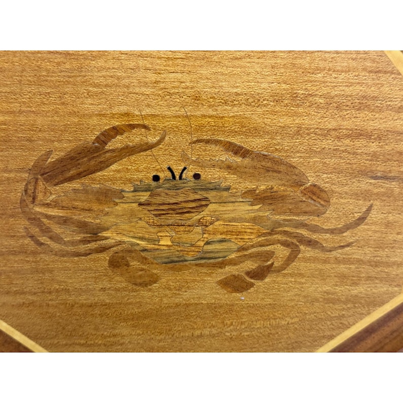 May include: Wooden inlay featuring a crab design. The crab is crafted from various wood tones, with a detailed body and claws. The background is a light-colored wood with a visible grain, and the overall design is geometric.