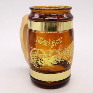 May include: A brown glass mug with a wooden handle. The mug has a gold band around the top and bottom. It features a yellow graphic of a roller coaster with the text "The Ragin' Cajun" printed on the side.