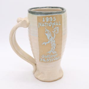 May include: Cream-colored ceramic mug with a large handle and a light blue rim. The mug features a tan rectangular panel with the text "1995 NATIONAL SHRIMP FESTIVAL" and a shrimp graphic in light blue.