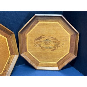 May include: Two octagonal wooden trays with a crab inlay. The trays are made of light and dark brown wood with a yellow border. The crab is made of various wood tones. The trays are likely used for serving or display.