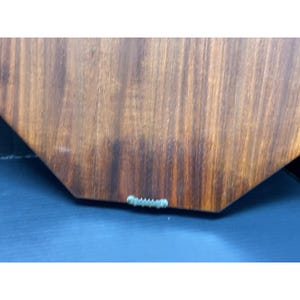 May include: Close-up of the back of a wooden wall art piece. The wood has a rich, warm brown tone with visible grain patterns. A metal sawtooth hanger is attached to the bottom edge for hanging. The art piece has a unique, geometric shape.