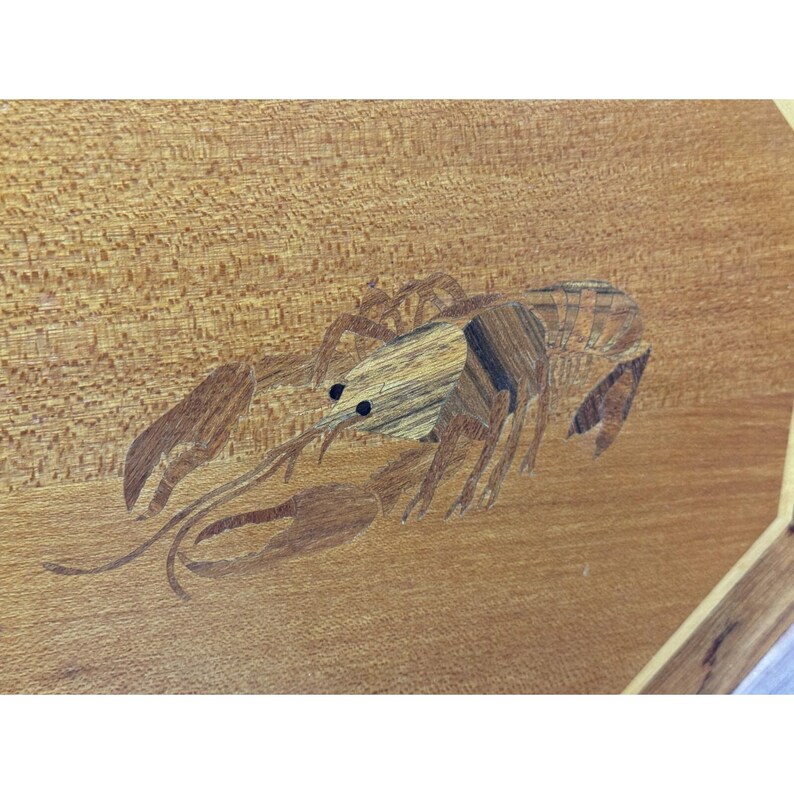 May include: A wooden surface with a detailed inlaid design of a lobster. The lobster is crafted from various wood tones, creating a textured and dimensional effect. The background is a light brown wood grain.