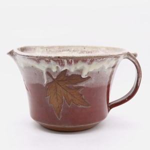 May include: A handmade ceramic mixing bowl with a handle and spout. The bowl is a deep red color with a brown leaf design and a white glaze around the rim. The bowl is perfect for mixing ingredients.