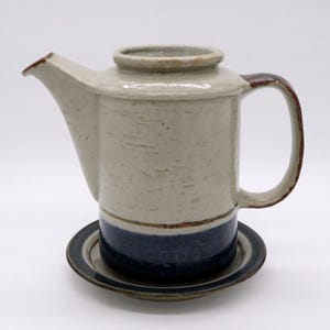 May include: A ceramic teapot with a matching saucer. The teapot is primarily beige with a dark brown handle and spout trim. A dark blue band encircles the base. The saucer is dark blue with a brown rim. The teapot is sitting on the saucer.
