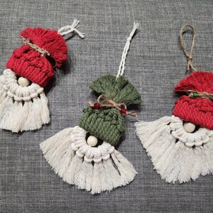 Könnte beinhalten: Drei handgefertigte Makramee-Weihnachtsornamente. Jedes Ornament hat eine rote oder grüne Mütze, einen weißen Bart und ein Gesicht aus Holzperlen. Die Ornamente sind mit Schnur oder Faden aufgehängt.