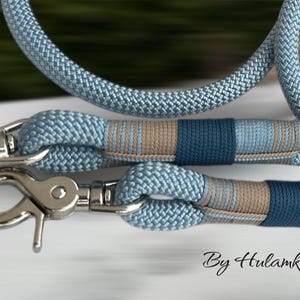May include: Close-up of a light blue dog leash with silver metal clasps. The leash features a woven rope design with sections of blue, tan, and navy blue. The text "By Hulamka" is visible in the bottom right corner.