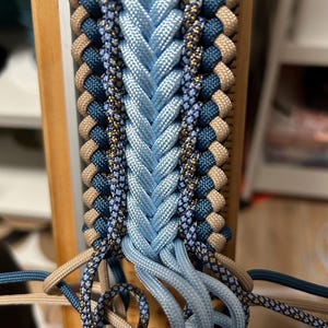 May include: Close-up of a braided craft project in progress. The design features a central braid of light blue cord, flanked by rows of navy, tan, and patterned cords. The cords are attached to a wooden frame, showcasing the intricate weaving process.