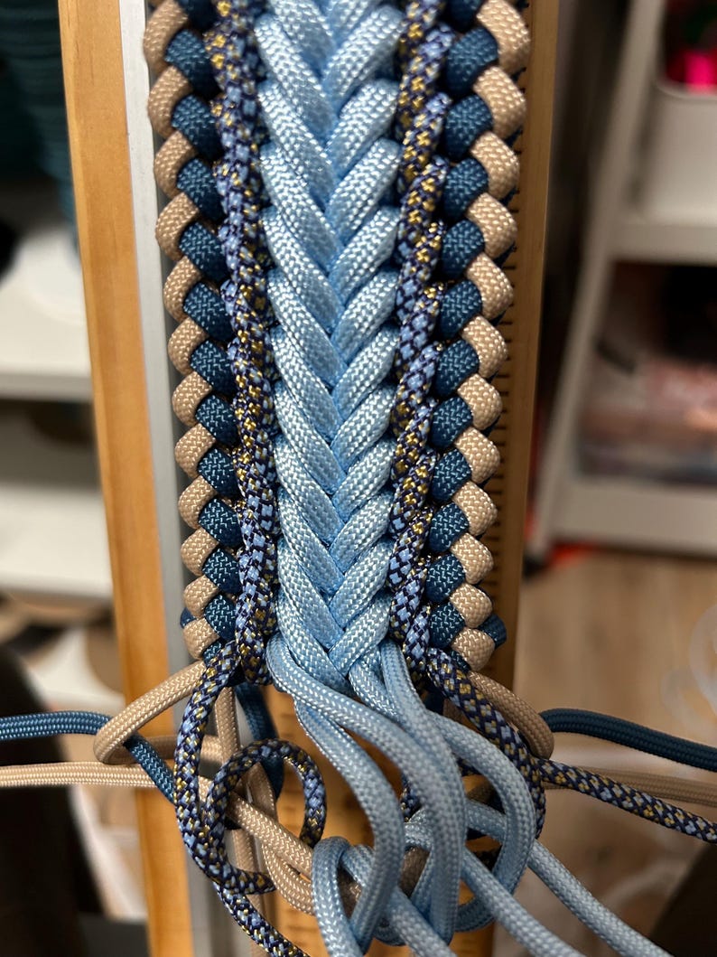 May include: Close-up of a braided craft project in progress. The design features a central braid of light blue cord, flanked by rows of navy, tan, and patterned cords. The cords are attached to a wooden frame, showcasing the intricate weaving process.