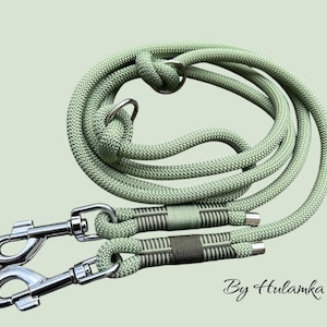 Può includere: Un guinzaglio per cani a doppia estremità di colore verde salvia con due moschettoni argentati e un anello argentato. Il guinzaglio è realizzato in corda intrecciata e presenta un motivo a righe. Il testo "By Hulamka" è visibile nell'angolo in basso a destra.