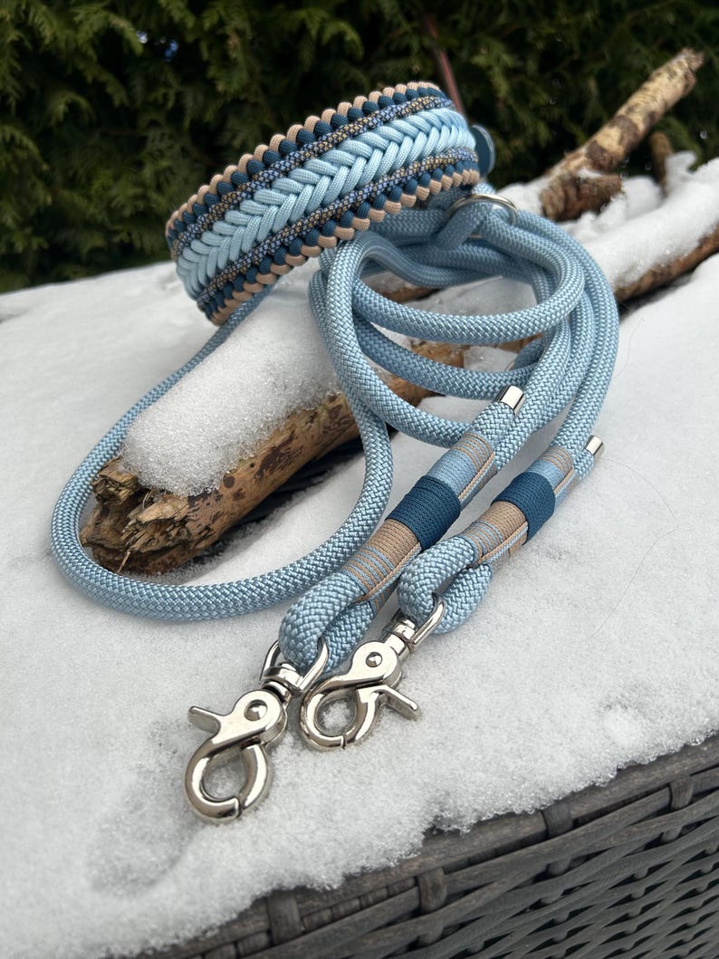 May include: A light blue dog collar and matching leash set. The collar features a braided design with light blue, tan, and navy blue accents. The leash has a light blue rope with silver-colored clasps.