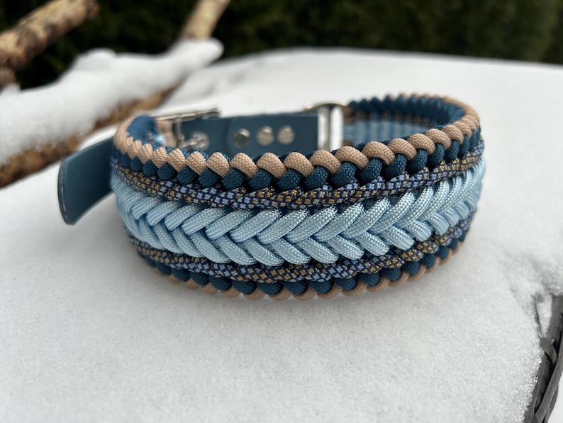 May include: A braided dog collar in shades of blue, tan, and navy. The collar features a light blue braided center section, with darker blue and tan accents. The collar is made of durable materials and has a silver buckle.