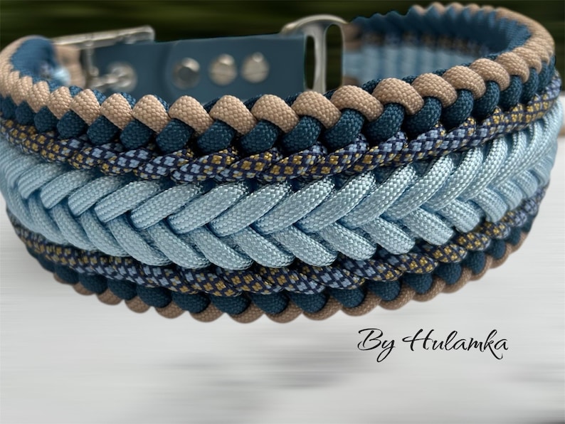 May include: A braided dog collar in shades of blue, tan, and navy. The collar features a light blue braided center, bordered by navy and tan rope. The collar has a silver-colored buckle and is labeled "By Hulamka".