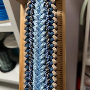 May include: A close-up of a braided paracord item in shades of blue, tan, and navy. The central braid is light blue, flanked by darker blue and tan braids. The item is attached to a wooden support with a metal ring.