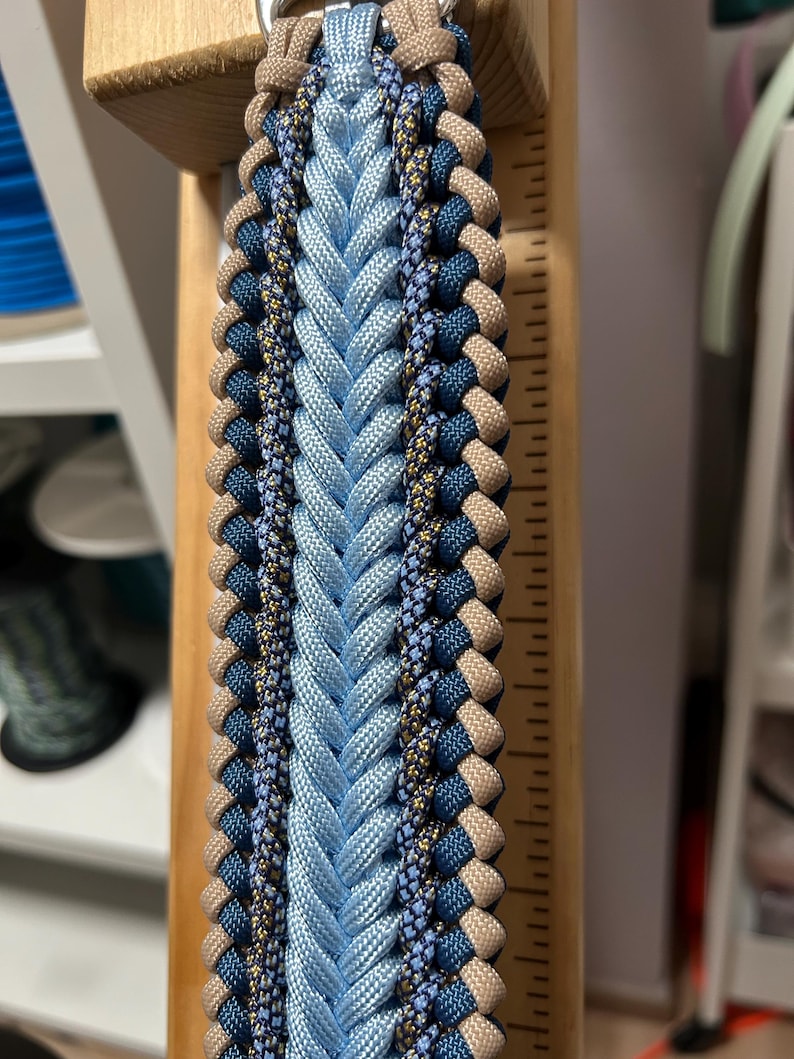 May include: A close-up of a braided paracord item in shades of blue, tan, and navy. The central braid is light blue, flanked by darker blue and tan braids. The item is attached to a wooden support with a metal ring.