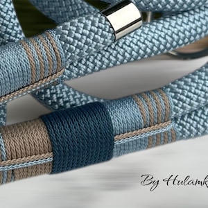 May include: Close-up of a light blue rope with decorative bands in shades of blue and brown. The rope has a woven texture and is accented with silver-colored metal clasps. The text "By Hulamka" is visible in the bottom right corner.