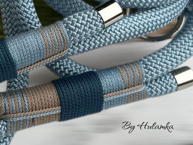 May include: Close-up of a light blue rope with decorative bands in shades of blue and brown. The rope has a woven texture and is accented with silver-colored metal clasps. The text "By Hulamka" is visible in the bottom right corner.