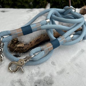 May include: A light blue dog leash with silver-colored metal clasps. The leash has a woven rope design with dark blue and tan accents. The leash is lying on a snowy surface.