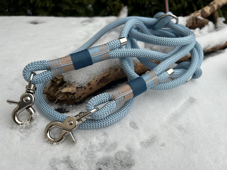 May include: A light blue dog leash with silver-colored metal clasps. The leash has a woven rope design with dark blue and tan accents. The leash is lying on a snowy surface.
