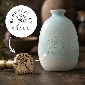 Puede incluir: Un jarrón de cerámica azul claro con cuello estrecho y acabado liso, que presenta un diseño estampado en relieve con el texto HANDMADE BY SHANN y un gráfico de diente de león. Al lado hay un sello de latón con el mismo logotipo circular.