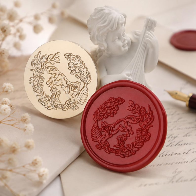 May include: A circular gold-toned metal wax seal stamp featuring an engraved fox surrounded by a floral wreath, paired with a matching red wax seal impression showing the same fox and floral design, with the letters MMG visible at the bottom.
