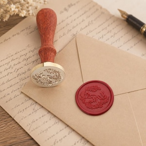 May include: A wax seal stamp with a turned wooden handle and a brass head featuring an engraved fox design. Beside it, a tan envelope displays a matching red wax seal impression of a fox surrounded by foliage, resting on aged parchment paper.
