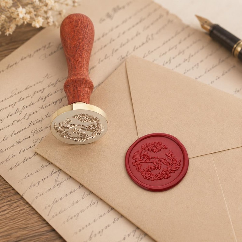May include: A wax seal stamp with a turned wooden handle and a brass head featuring an engraved fox design. Beside it, a tan envelope displays a matching red wax seal impression of a fox surrounded by foliage, resting on aged parchment paper.