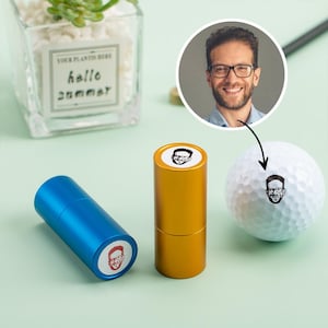 May include: A golf ball with a personalized face stamp next to two cylindrical stampers, one blue and one gold. The stampers and golf ball feature a black and white image of a person's face. The background is a light green color.