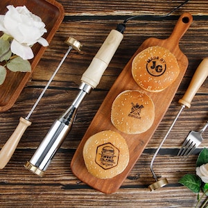 Puede incluir: Una tabla de cortar de madera con tres bollos de hamburguesa con semillas de sésamo marcados con logotipos. Un hierro de marcar con mango de madera y cabeza de metal está al lado de los bollos. También se ven dos hierros de marcar y un tenedor.