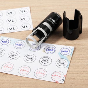 May include: A black and white self-inking stamp with the text "trodat printy 4612" next to its black holder. The stamp is next to several printed labels with the text "VL", "K&P", "Mr.P", and "Hana".