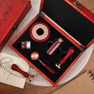 May include: A wax seal kit in a red wooden box containing two red-handled metal stamps, a metal melting spoon with a red handle, a small glass jar of red wax beads, and a round red wax warmer. One stamp is placed outside the open box.