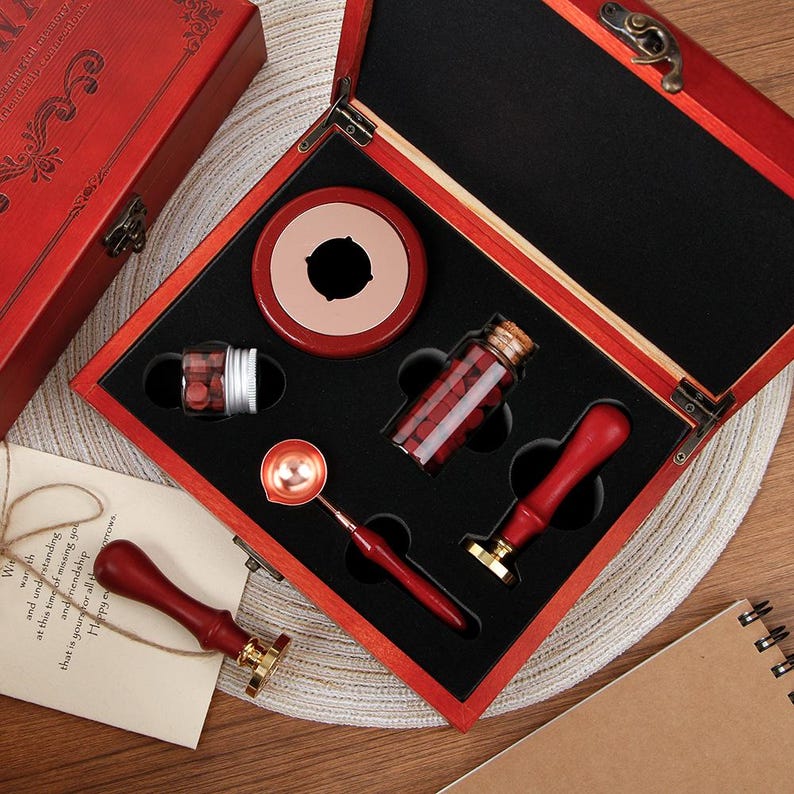 May include: A wax seal kit in a red wooden box containing two red-handled metal stamps, a metal melting spoon with a red handle, a small glass jar of red wax beads, and a round red wax warmer. One stamp is placed outside the open box.