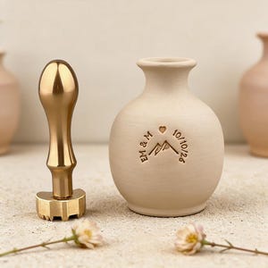 May include: A gold-colored wax seal stamp and a small, beige ceramic vase. The vase has a mountain and heart design with the initials "EM & M" and the date "10/10/26" engraved. Two small flowers are in the foreground.