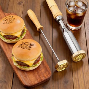 May include: Two cheeseburgers on a wooden cutting board, branded with "Chris BBQ" and "Christopher" logos. A metal branding iron with a wooden handle and a glass of cola with ice cubes are also visible.