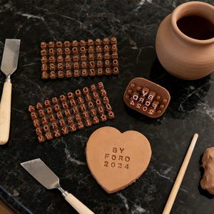 以下が含まれることがあります： 暗い表面に、茶色の粘土道具とスタンプが並んでいます。道具には、2つのこて、木のダボ、小さな粘土のポットが含まれています。スタンプには文字と「BY FORD 2024」の文字がハート型の粘土片に刻まれています。